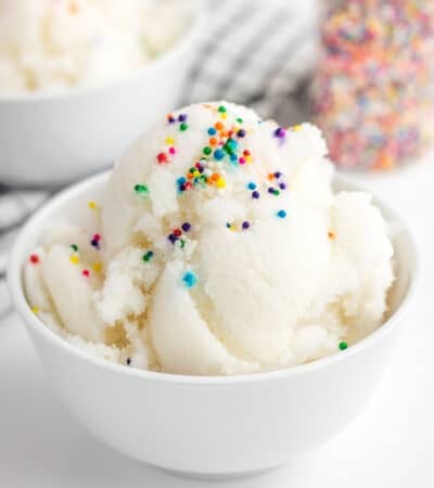 Vanilla ice cream in a white bowl with rainbow sprinkles. Another bowl of ice cream and a bottle of sprinkles can be seen in the background.