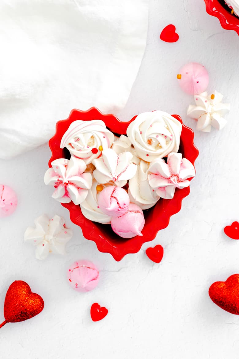Pink and white meringue cookies decorated with Valentine sprinkles and in a red heart-shaped bowl