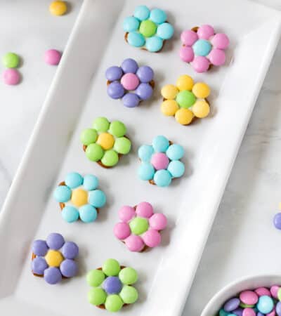 10 pastel-colored flowers made from M&Ms and pretzels displayed on a white serving plate with additional M&Ms sprinkled around the tabletop
