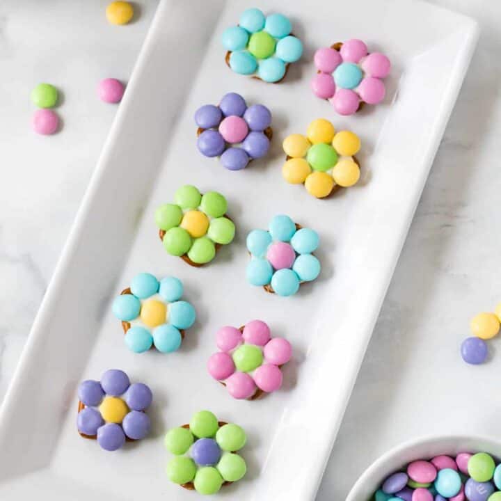 10 pastel-colored flowers made from M&Ms and pretzels displayed on a white serving plate with additional M&Ms sprinkled around the tabletop