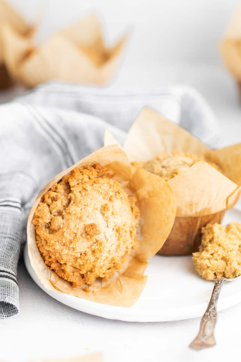 Wrapped crumb cake muffin, spoonful of crumb topping, and an unwrapped muffin on a white plate