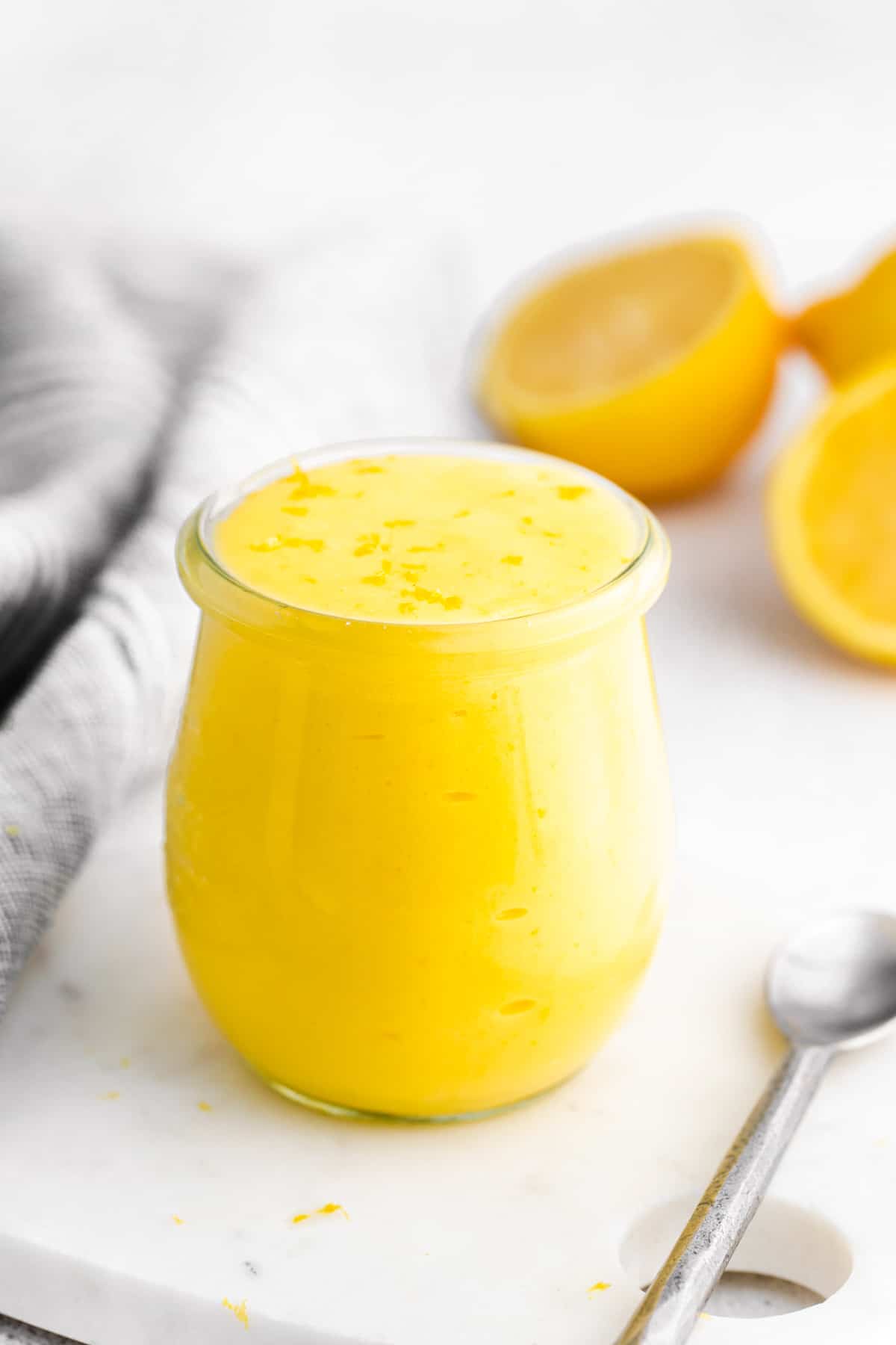 Lemon curd in a glass jar, with lemon halves and a silver spoon next to it