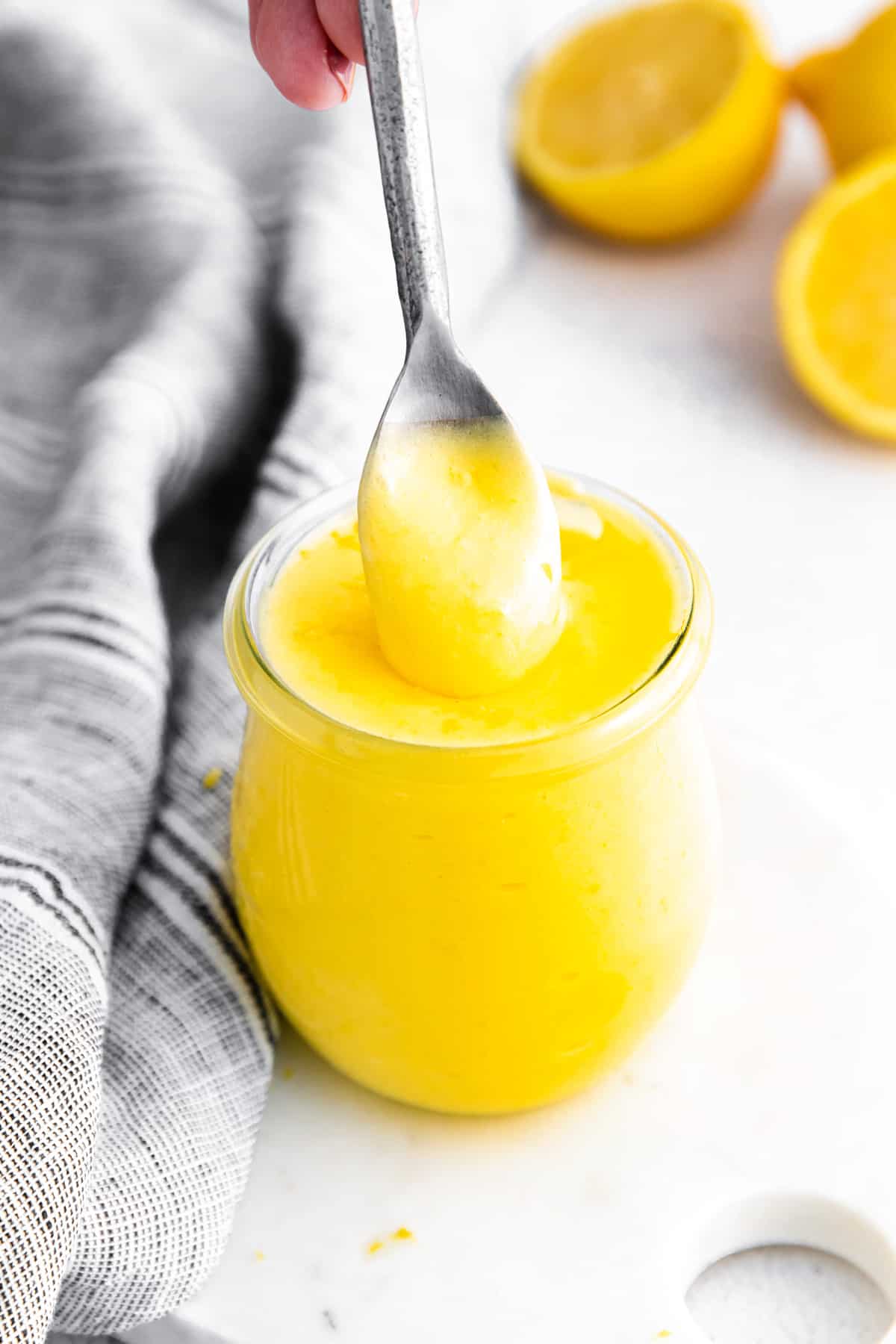 small glass jar of bright yellow lemon curd; a silver spoon is pulling out a spoonful from the top to show the creamy texture. Lemon halves and grey linen in background.