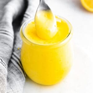 small glass jar of bright yellow lemon curd; a silver spoon is pulling out a spoonful from the top to show the creamy texture. Lemon halves and grey linen in background.