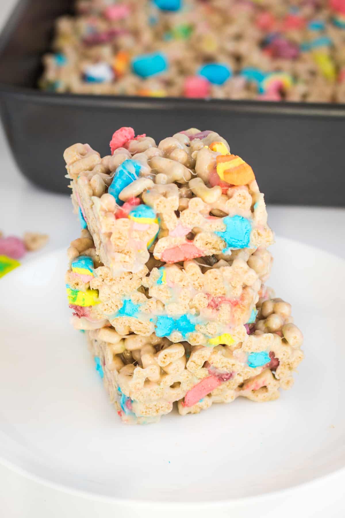 Three lucky charms marshmallow cereal bars cut into squares and stacked on top of one another on a white plate. A baking pan with uncut treats is behind them.