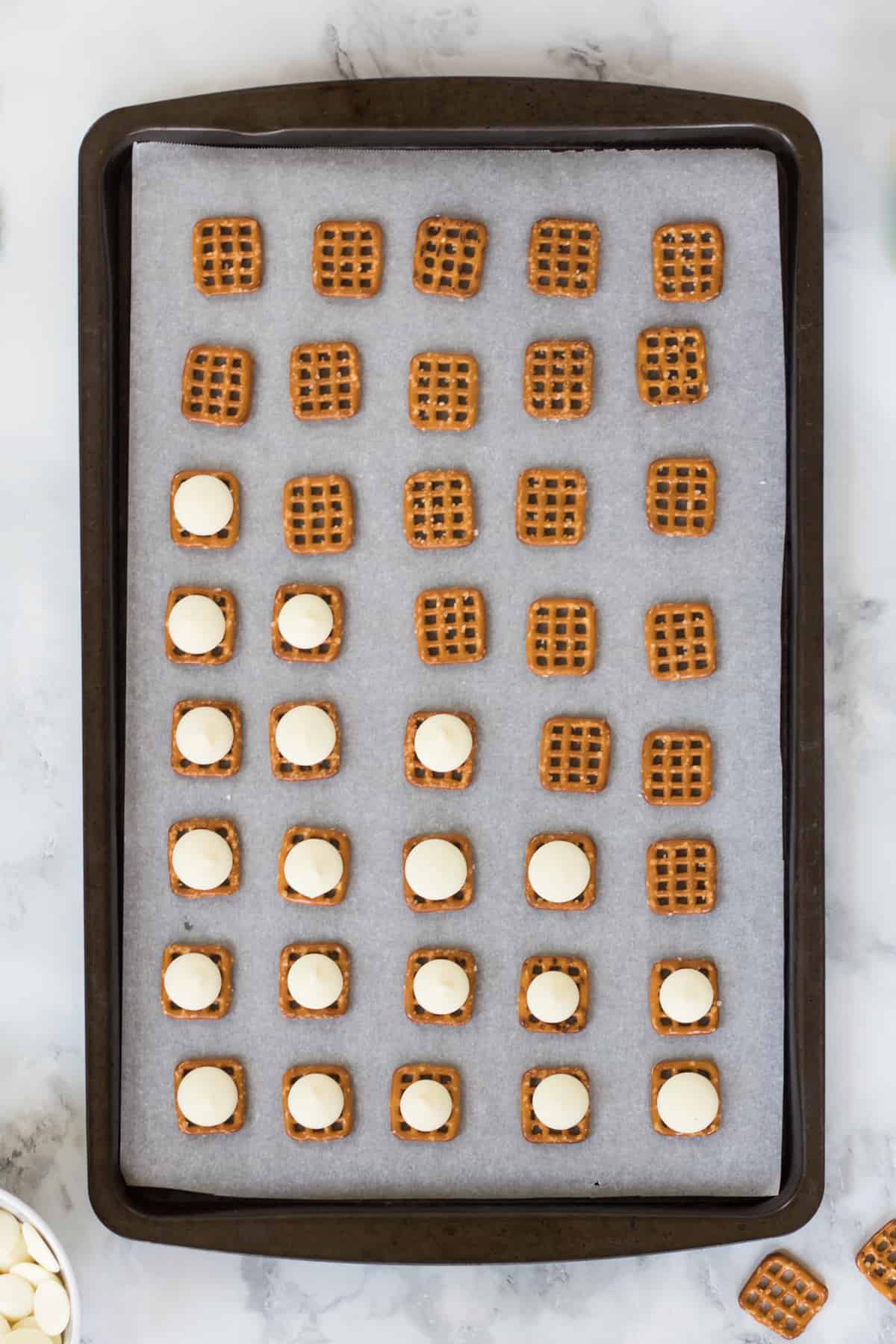 Baking sheet with rows of square pretzels. Half of the pretzels are topped with white chocolate melting wafers.