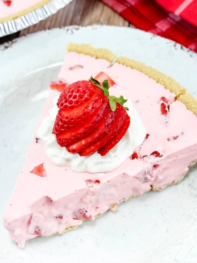 Slice of creamy pink pie with graham cracker crust and pieces of fresh berries in it. Pie is topped with whipped topping and a sliced strawberry for garnish