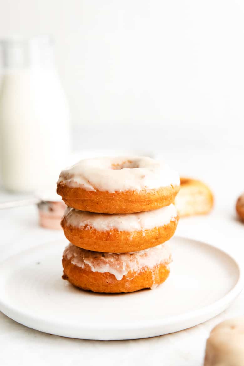 Three glazed doughnuts stacked on top of one another, on a white plate. A glass milk jug filled with milk is in background
