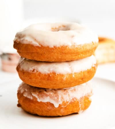 Three glazed doughnuts stacked on top of one another, set atop a white plate