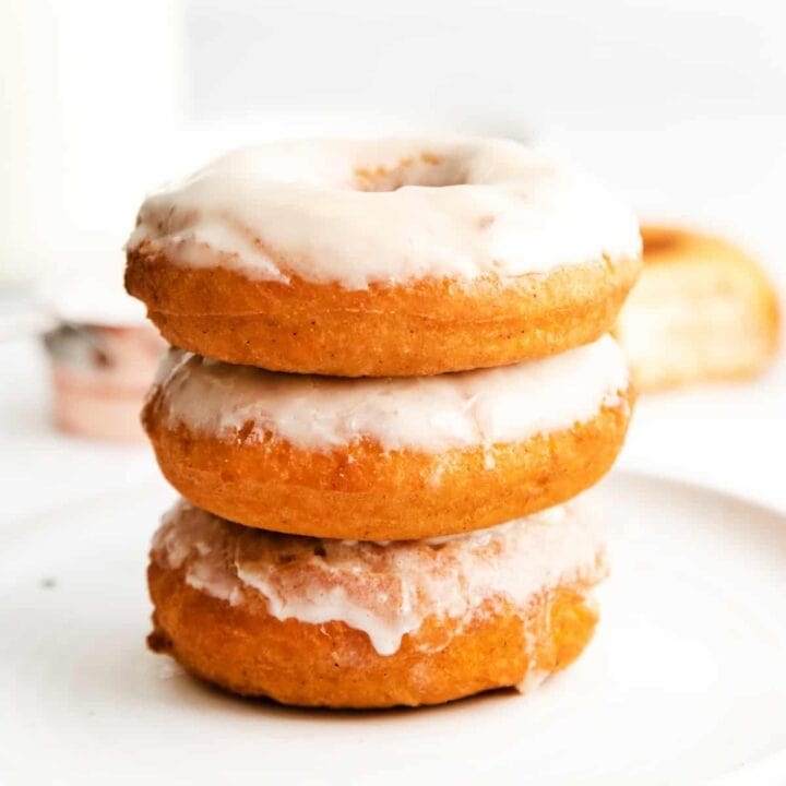 Three glazed doughnuts stacked on top of one another, set atop a white plate