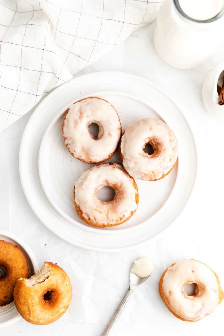 Three glazed cake doughnuts on a white plate surrounded by a white linen towel, glass jug of milk, a spoon, and additional doughnuts, some un-glazed
