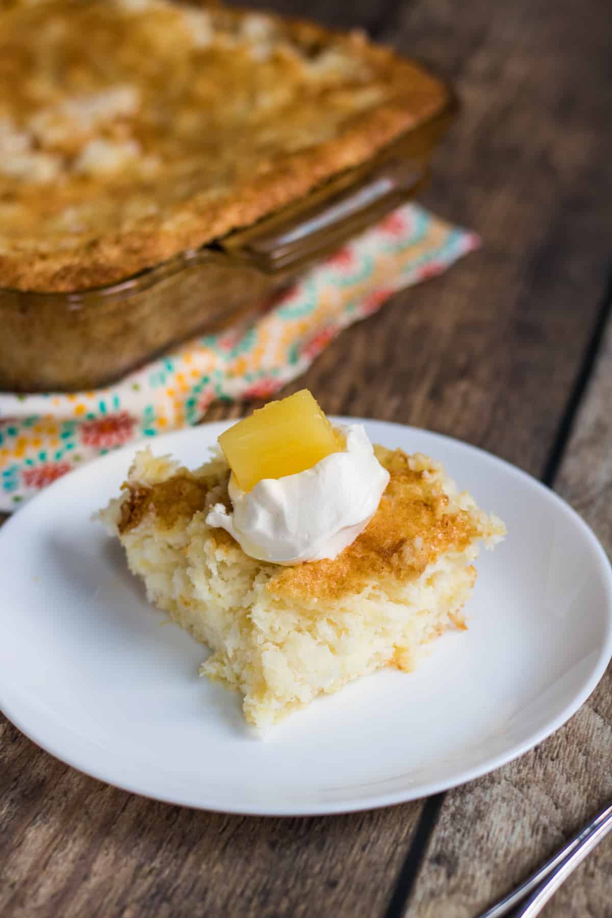 Square slice of angel food cake with golden brown top on a white plate. Cake is topped with a dollop of whipped topping and a chunk of pineapple . Rectangular baking dish with cake in background.