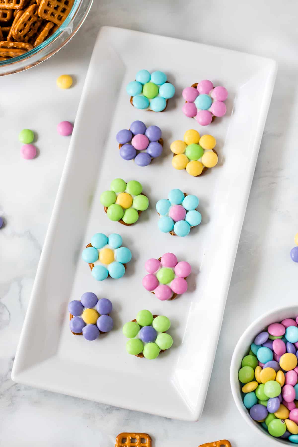 10 pastel-colored flowers made from M&Ms and pretzels on a white serving platter. A bowl of pretzel snaps and a bowl of M&Ms are next to the platter.