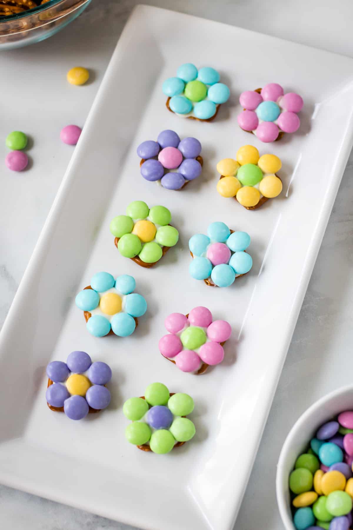 Pastel-colored flowers made of M&Ms and pretzels on a white serving plate. Bowls of more M&Ms and pretzels are next to platter.