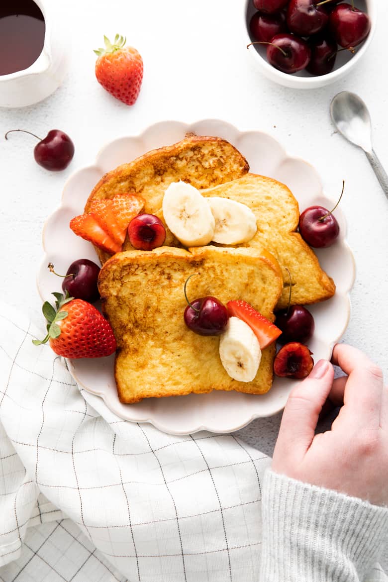 Brioche french toast with banana slices and fresh berries on a white plate with a woman's hand reaching out and placing the plate on the table. Maple syrup, cherries, strawberries, a spoon, and a white linen napkin are surrounding plate