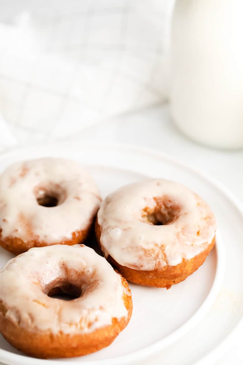 Three glazed donuts on a white plate with a glass container of milk next to them