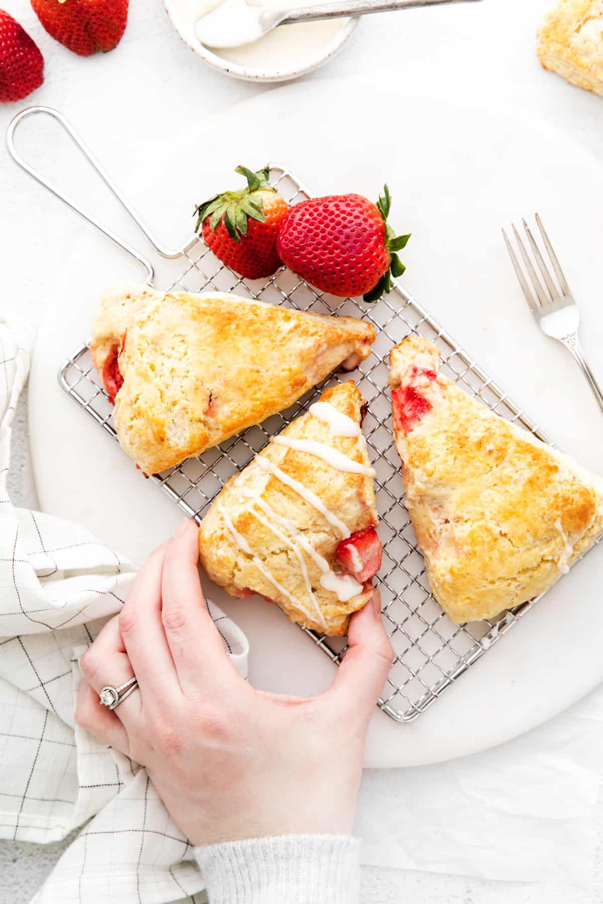 Woman's hand reaching to take a homemade strawberry scone. Scone is on wire holder with 2 other scones and 2 fresh strawberries. 