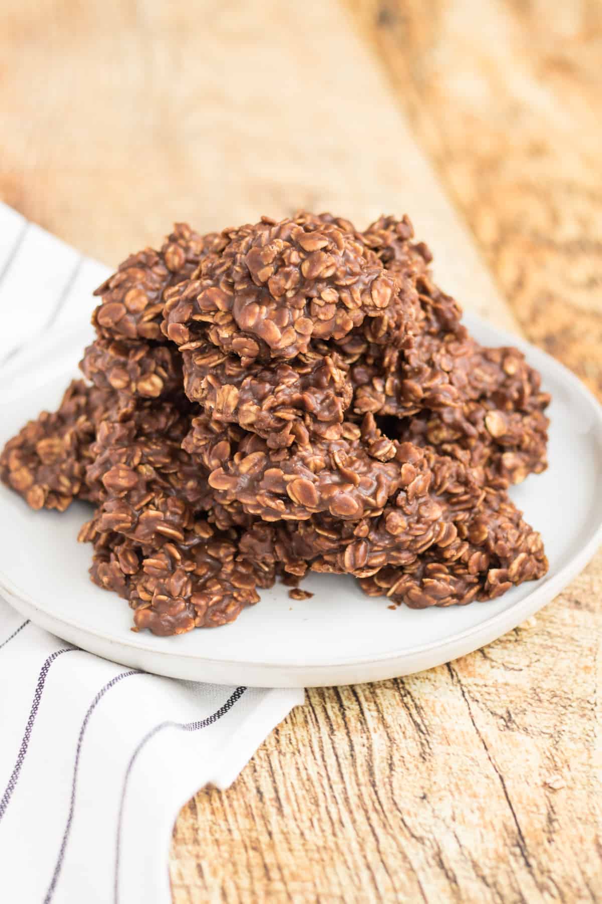 Chocolate oatmeal cookies piled on a white plate with a white napkin with grey stripe under it on a light wood table