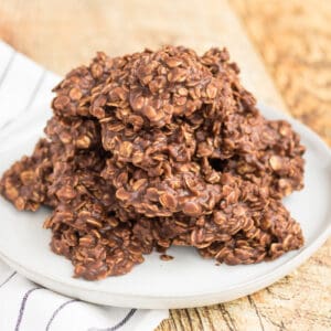 Chocolate oatmeal cookies piled on a white plate with a white napkin with grey stripe under it on a light wood table