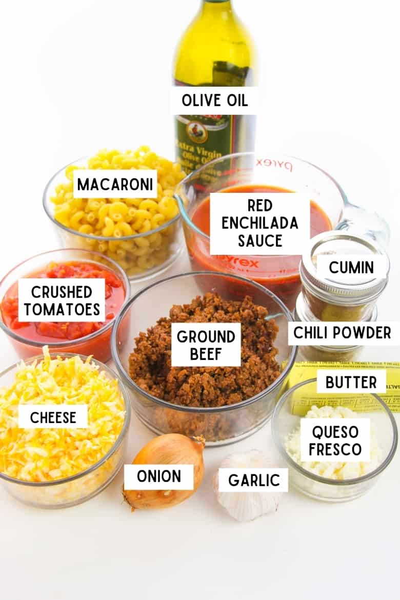 labeled recipe ingredients in glass bowls: macaroni, red enchilada sauce, cumin, chili powder, queso fresco, shredded cheese, crushed tomatoes, and ground beef. Also, a stick of butter, onion, garlic, and bottle of olive oil