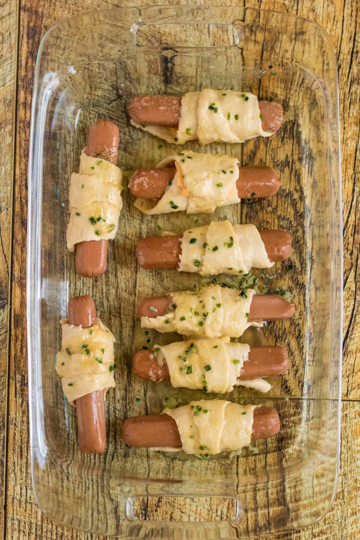 Unbaked crescent-wrapped hot dogs arranged in glass baking dish and topped with garlic and chive butter