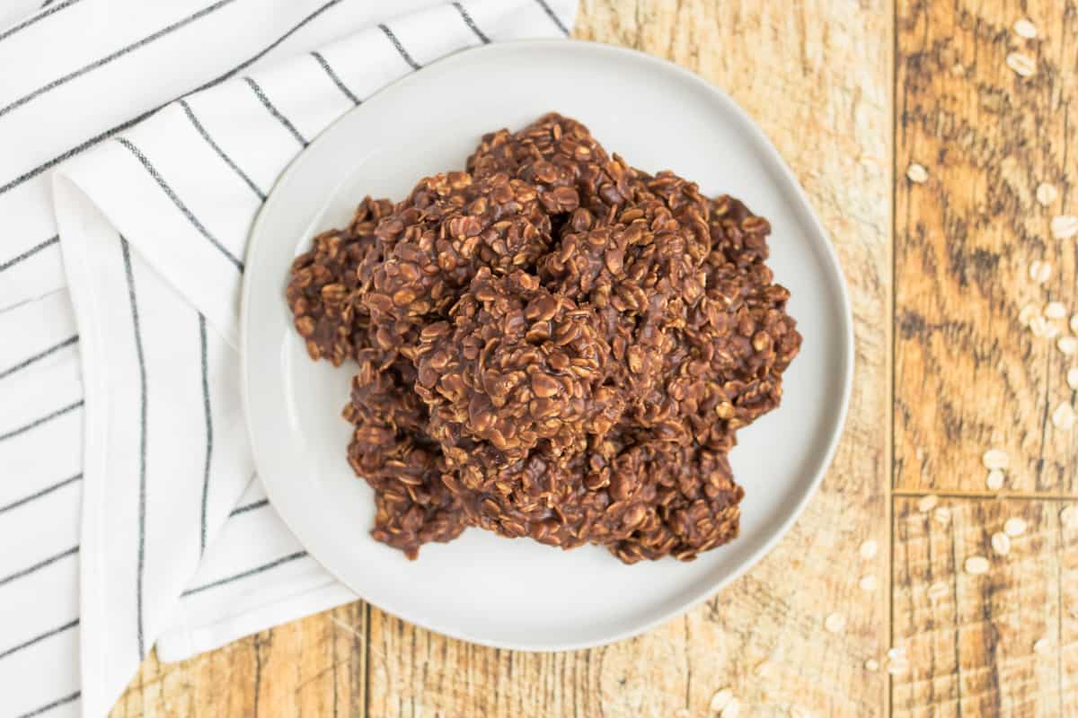 Chocolate no bake cookies piled on a white plate with a white napkin with grey stripe under it on a light wood table