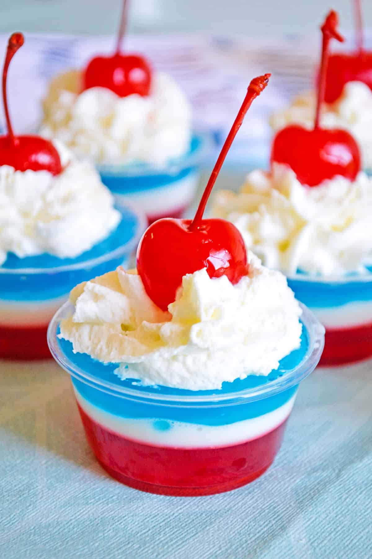 Layers of red, white, and blue gelatin topped with whipped cream and a maraschino cherry in a small clear plastic cup