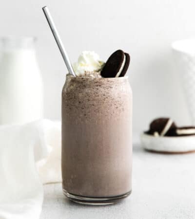 OREO cookie milkshake in tall glass with whipped cream, an oreo cookie, and straw sticking out of the top. Pitcher of milk and plate of cookies in background.