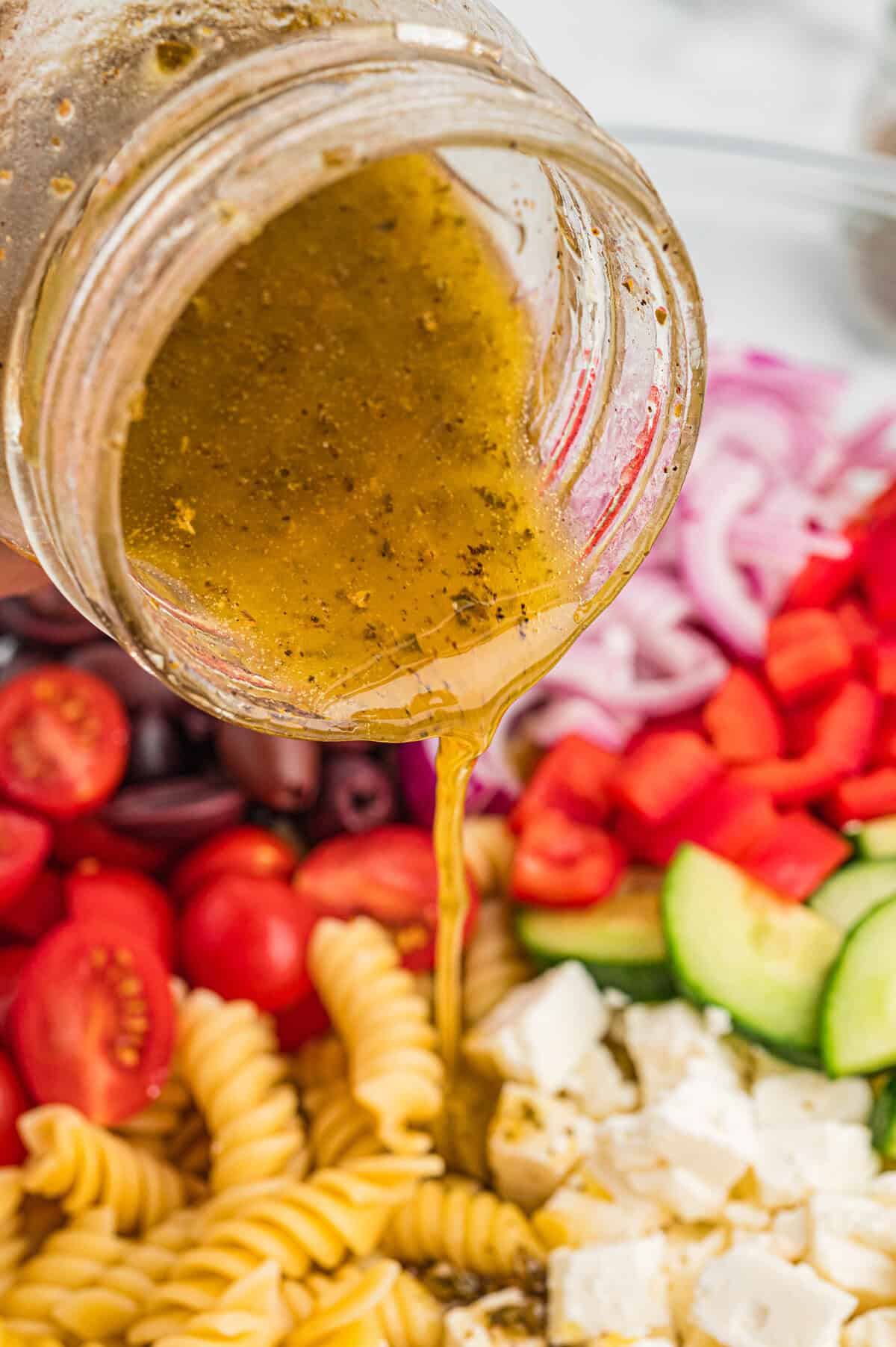 A jar of homemade vinaigrette dressing is being poured over Greek pasta salad ingredients including rotini, cherry tomatoes, kalamata olives, red onion, red bell pepper, cucumber, and feta cheese.