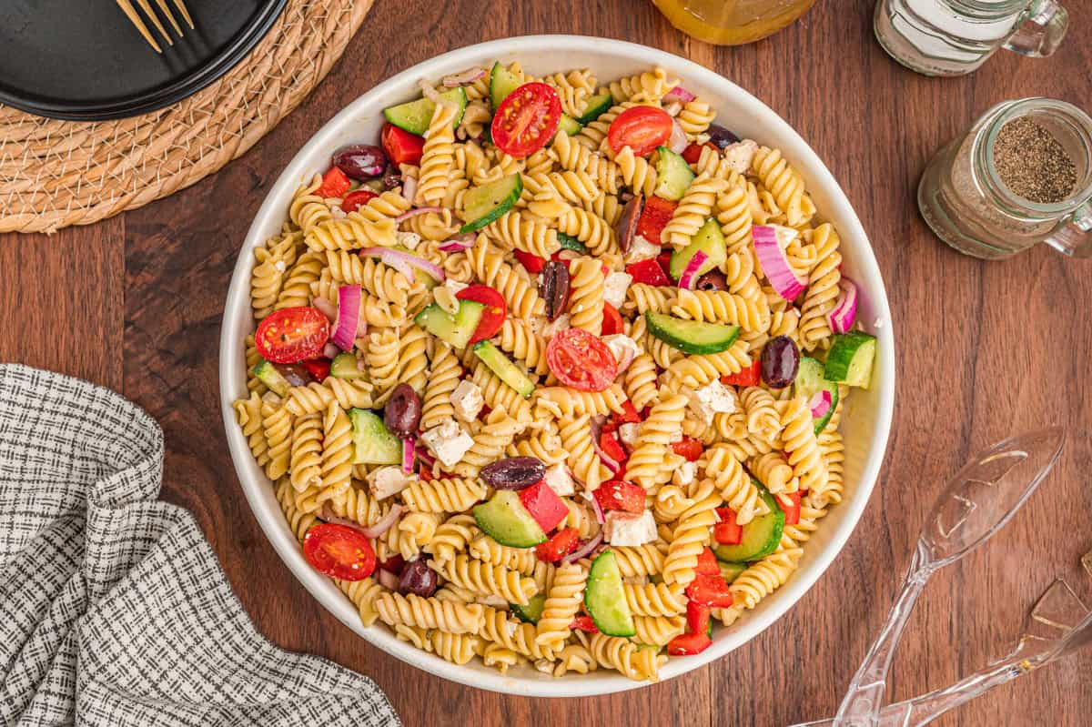A white bowl filled with Greek pasta salad on a wooden surface, containing rotini pasta, cherry tomatoes, cucumber slices, kalamata olives, feta cheese, red onion, and red bell pepper.