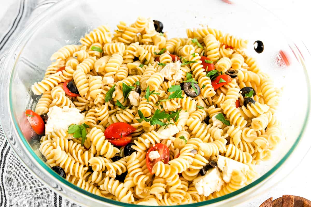 Large glass bowl full of a pasta salad with halved cherry tomatoes, crumbled feta cheese, sliced black olives, and a sprinkle of parsley.