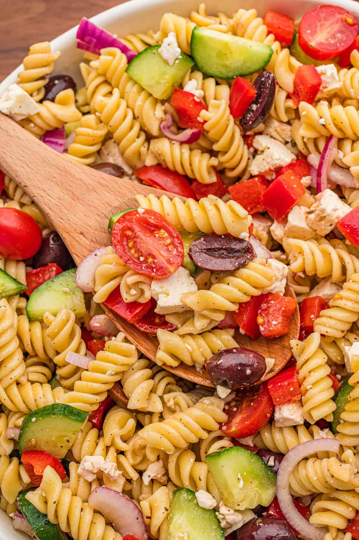 Greek pasta salad with rotini, halved cherry tomatoes, cucumber slices, red bell pepper, kalamata olives, feta cheese, red onion, and homemade dressing served on a wooden spoon.