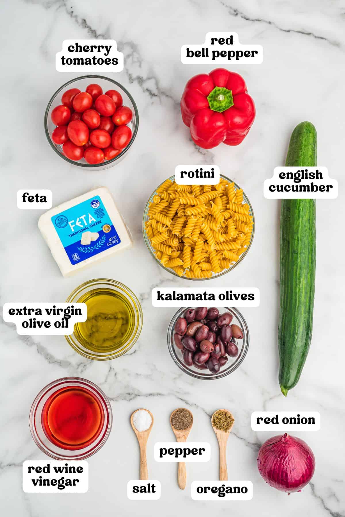 Overhead image of ingredients for Greek pasta salad, each labeled: cherry tomatoes, red bell pepper, English cucumber, red onion, feta, rotini, kalamata olives, extra virgin olive oil, red wine vinegar, salt, pepper, and dried oregano.