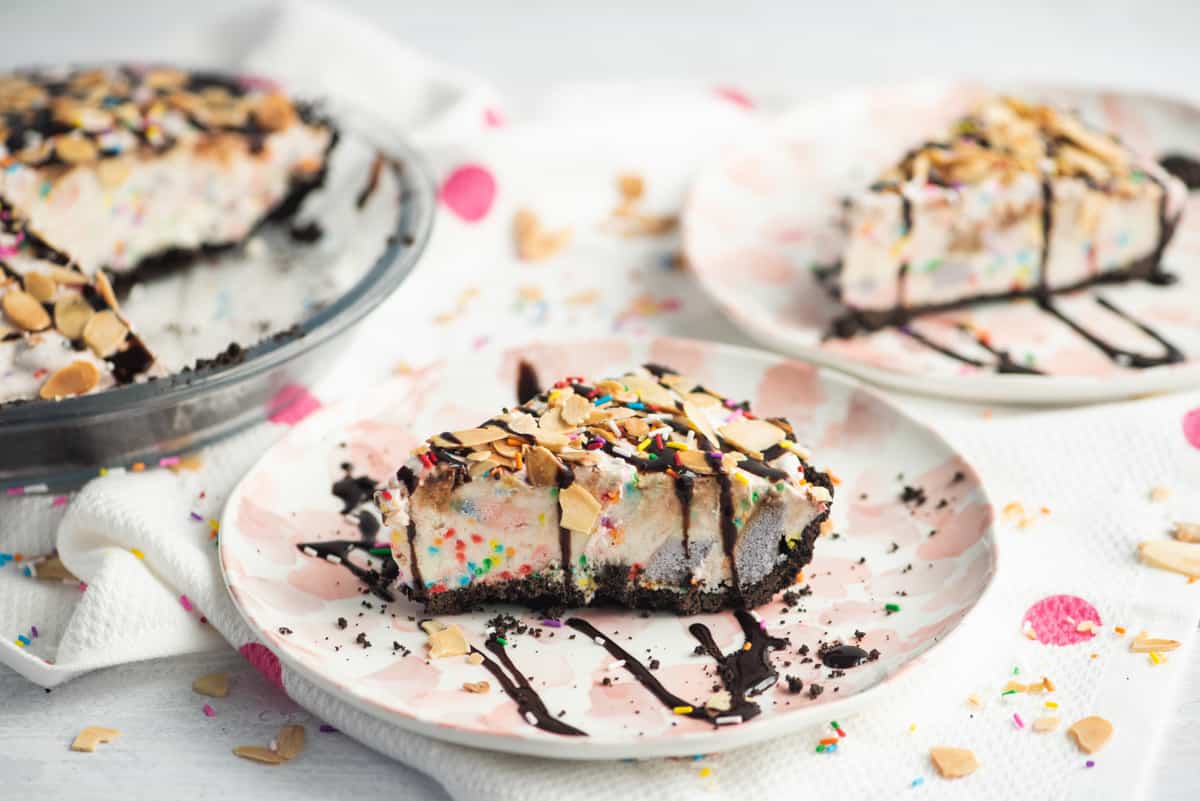 A slice o ice cream pie with chocolate crust and rainbow sprinkles. Pie is topped with drizzle of chocolate syrup and sliced almonds. Another slice and the remainder of the pie can be seen in the background.