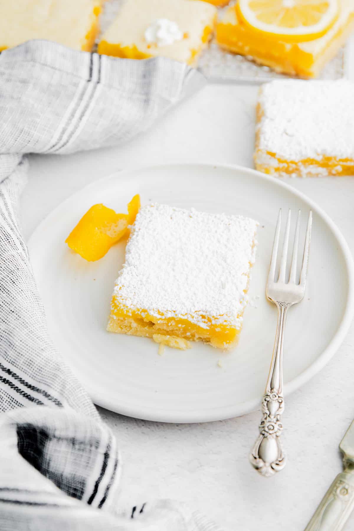 Lemon bar topped with powdered sugar on a white plate with a silver fork and garnish of lemon peel.