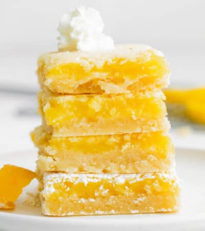 four bright yellow lemon Bars stacked on top of one another on a white plate.