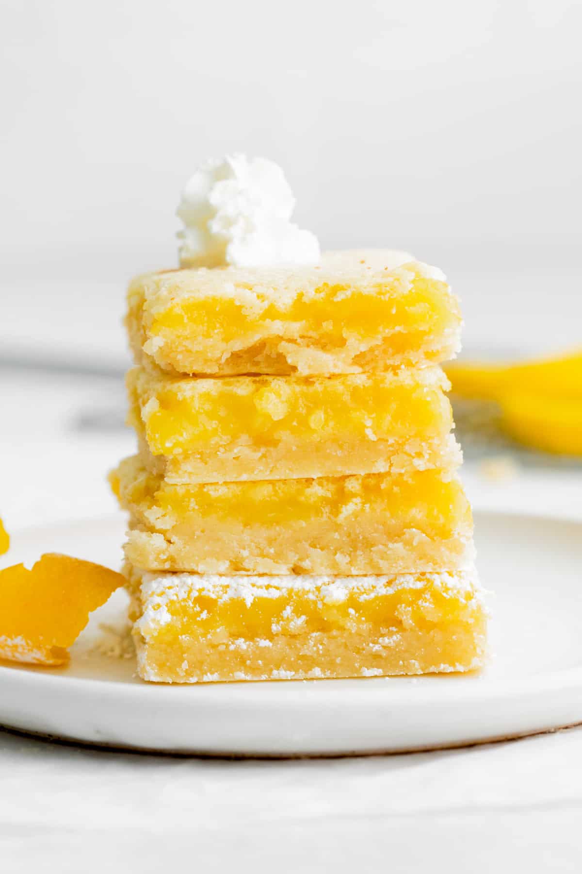 Four square lemon bars stacked on a white plate and topped with a small dollop of whipped cream