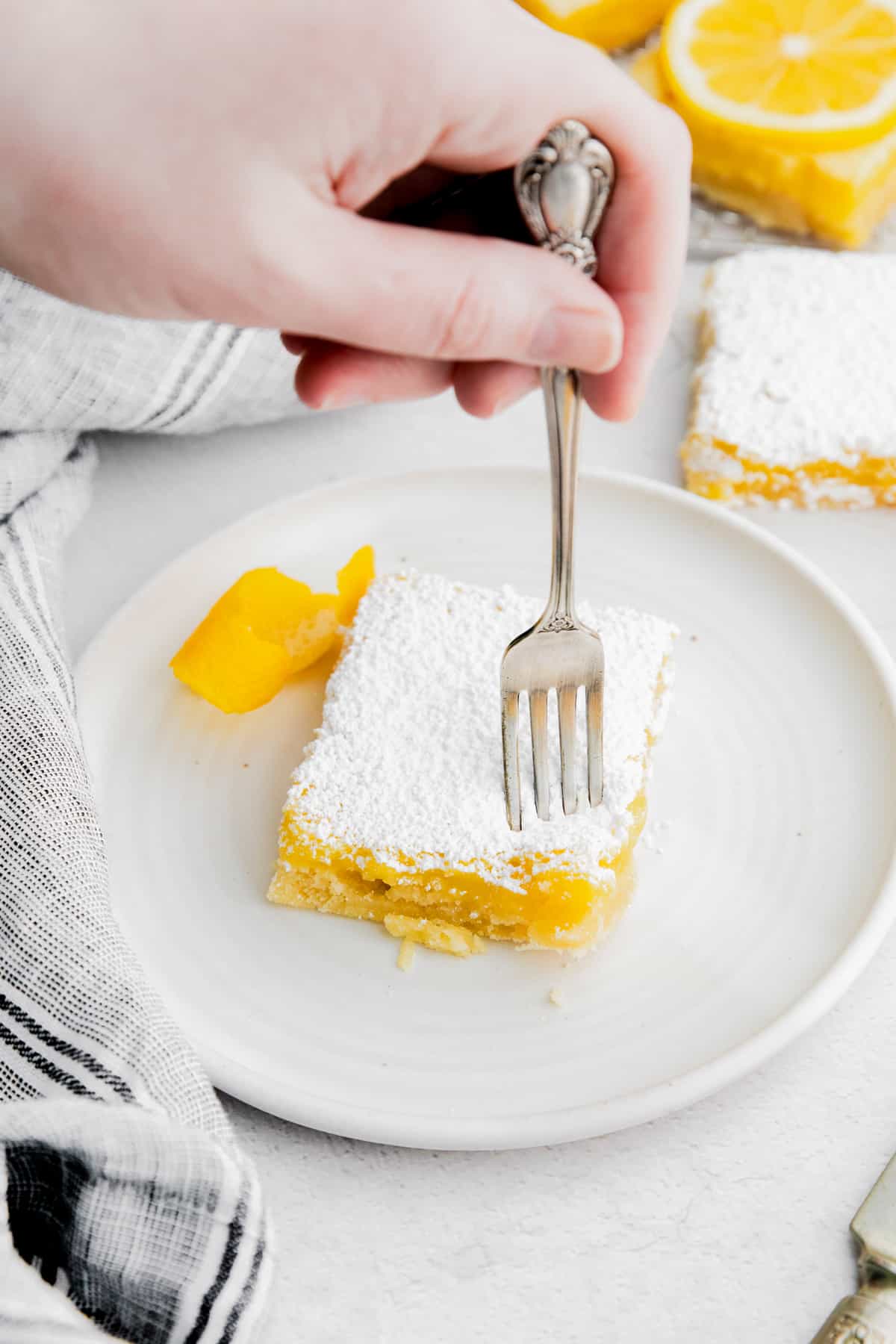 Hand holding a fork talking a piece out of a Lemon Bar topped with powdered sugar, served on a white plate with a garnish of lemon peel. 
