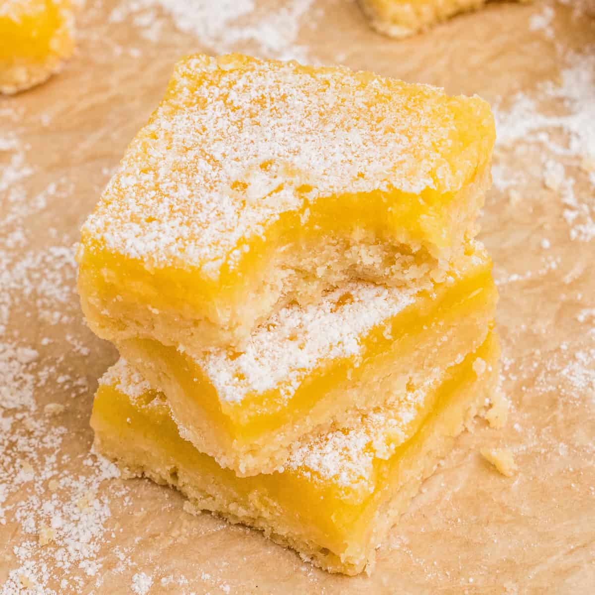 3 square lemon bars with shortbread crust and dusting of powdered sugar stacked. The top bar has a bite taken out of it.