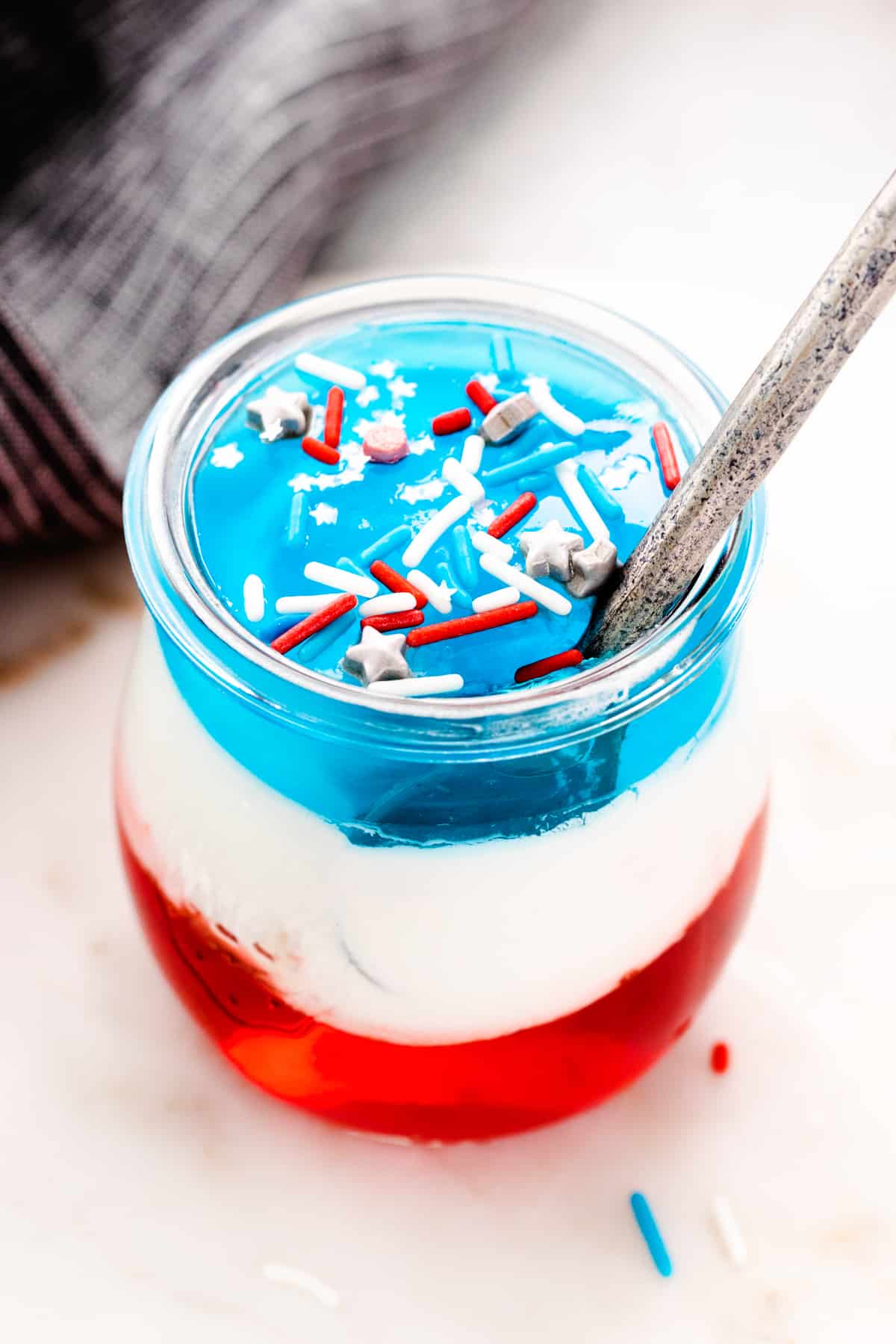 Individual size red, white, and blue layered jello cup with sprinkles and a silver spoon sticking out of the cup.