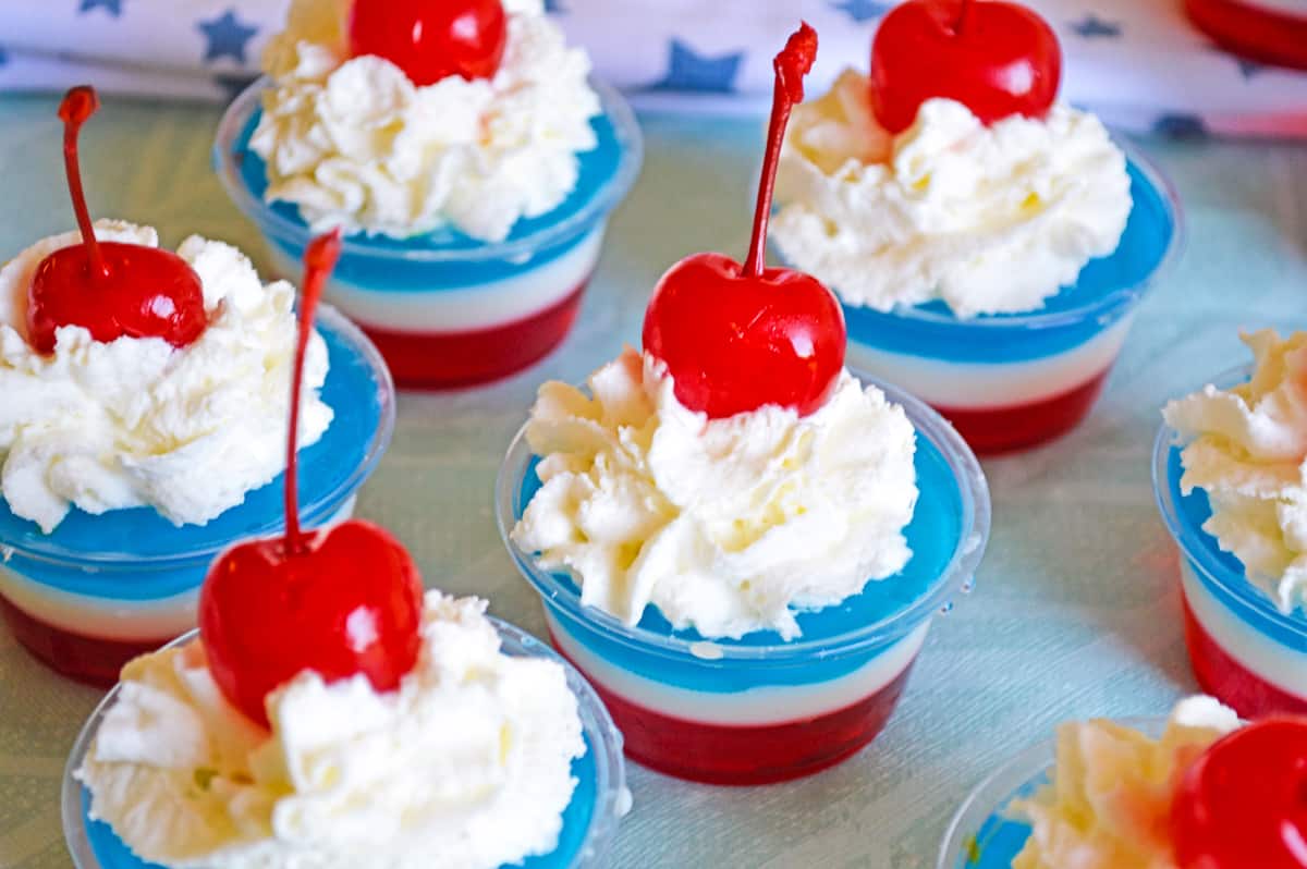 several red, white, and blue jello shots topped with whipped cream and cherries in small disposable plastic cups.