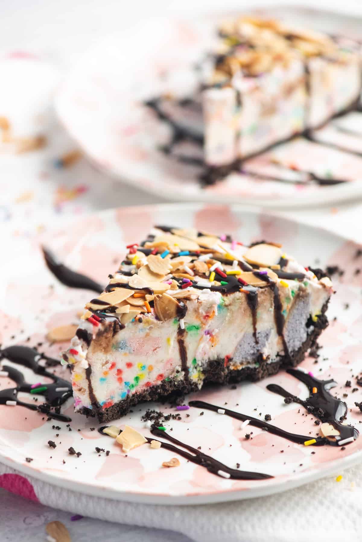 A slice of vanilla ice cream pie with chocolate crust topped with chocolate sauce, rainbow sprinkles, and sliced almonds. Chunks of rainbow cake and sprinkles can be seen inside the vanilla ice cream pie.