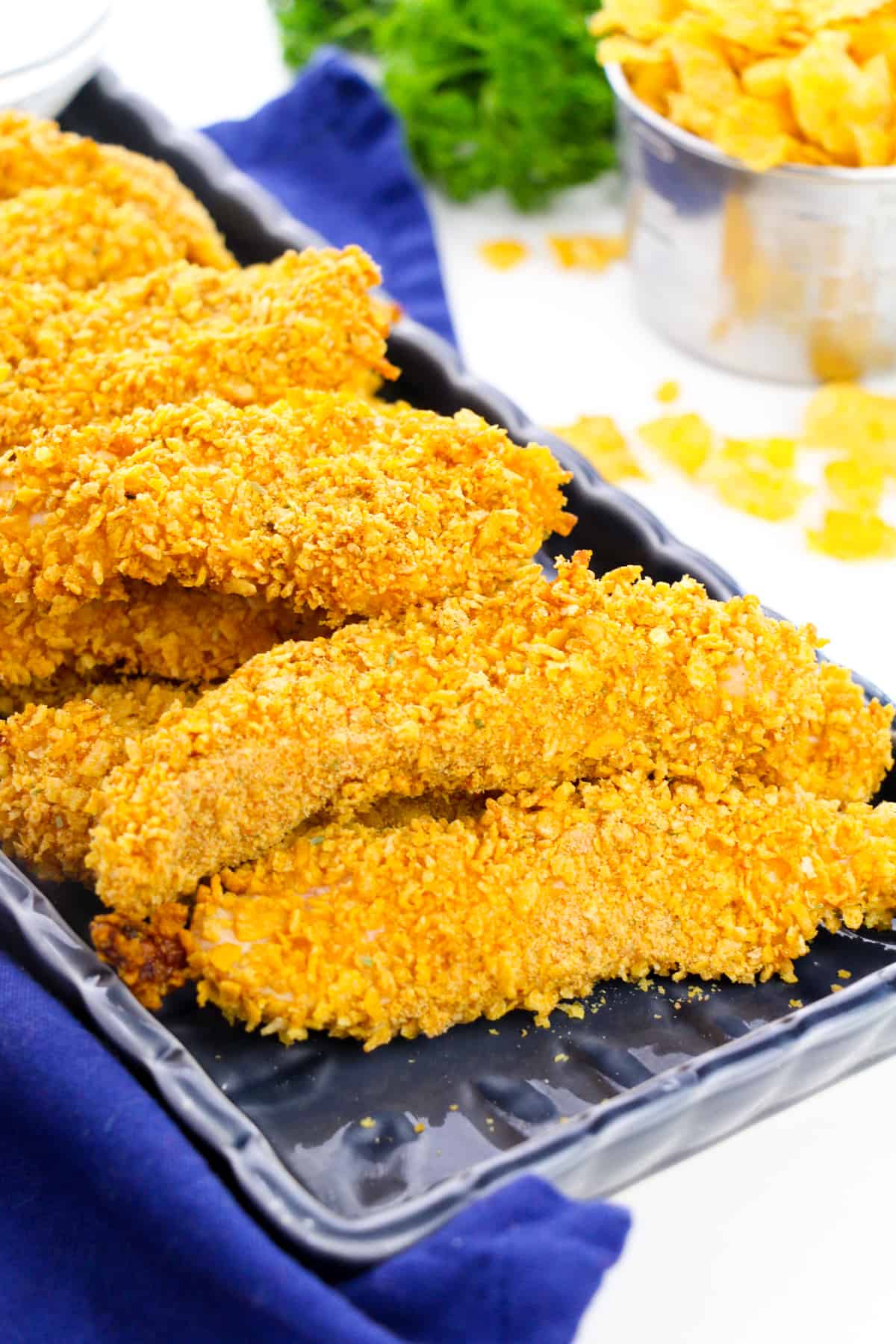 Cornflake chicken tenders on black plate with cornflakes and herbs in background