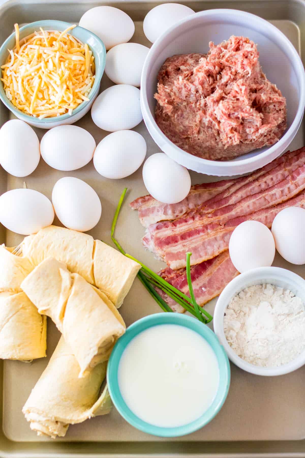 On a sheet pan: bowl of shredded cheese, 12 eggs, uncooked breakfast sausage, bacon, milk, flour, crescent roll dough, and fresh chives.