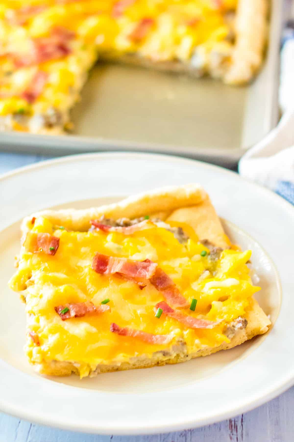 Square slice of sheet pan breakfast pizza on white plate, with remaining pizza in sheet pan in background.