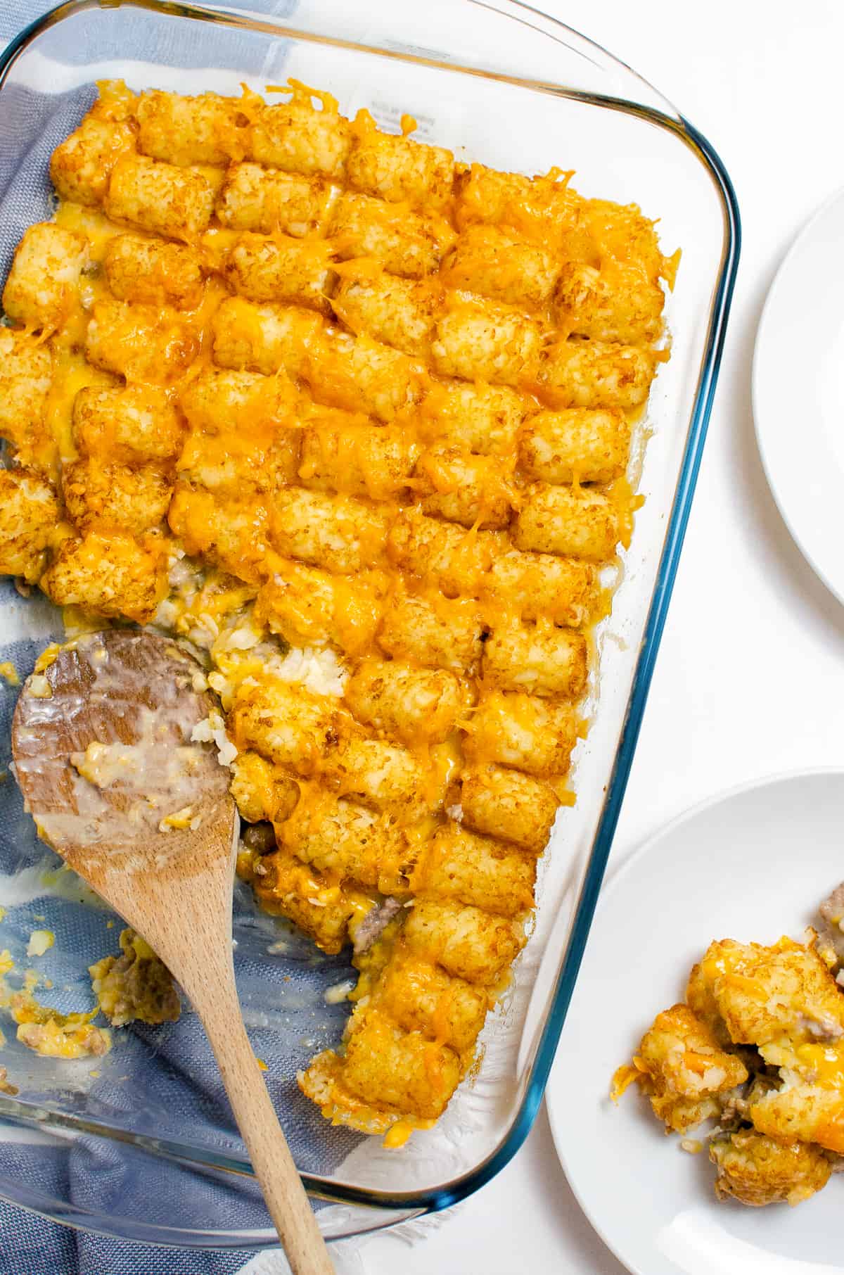 Tater tot casserole with ground beef and melted cheese in a 9 x 13 dish. A wooden spoon in resting in the half-empty dish and a serving of the casserole can be seen on a white plate halfway out of the frame