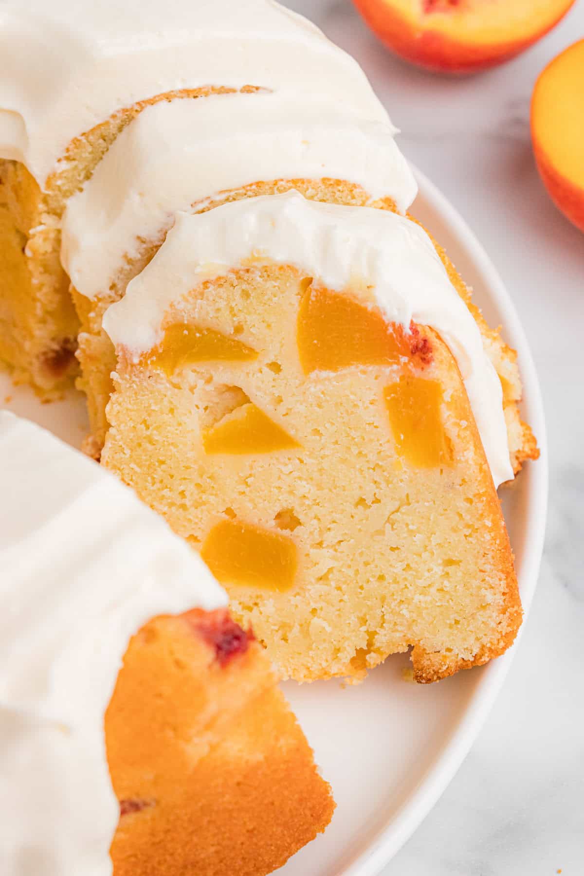 A sliced peach pound cake arranged on a white plate. The peach cake is topped with cream cheese icing, and the interior reveals multiple peach chunks.