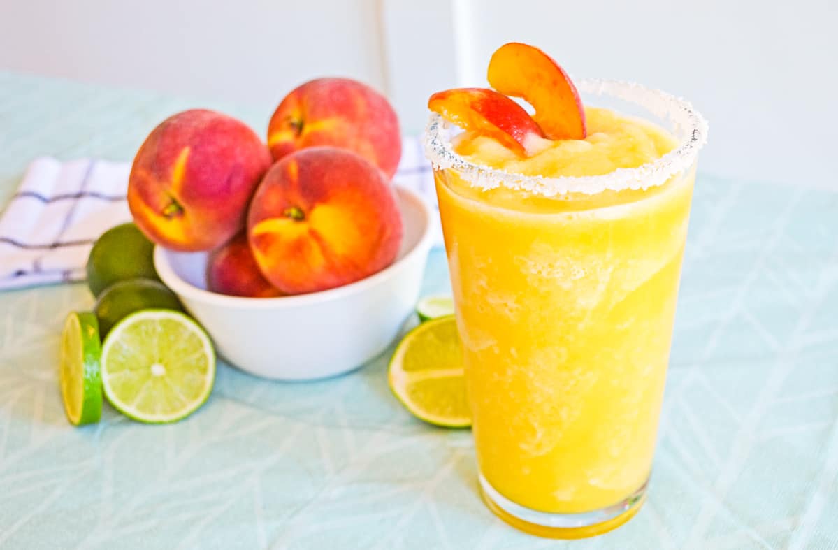 Frozen Peach margarita in glass with salt rim and garnished with fresh peaches. Bowl of fresh peaches and limes are behind the glass