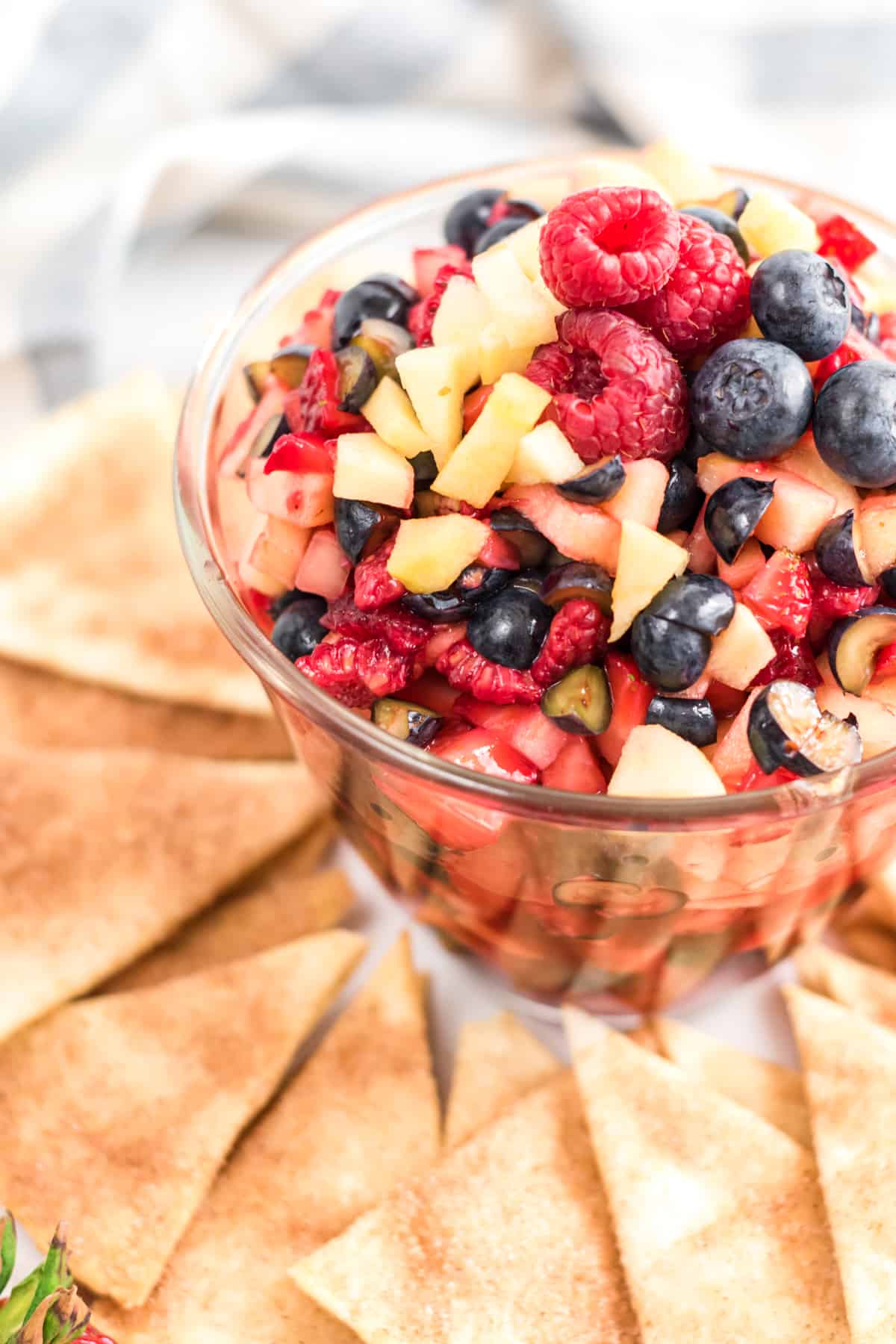 clear bowl of fruit salsa made with raspberries, strawberries, blueberries, and apples. The bowl is surrounded by cinnamon sugar tortilla chips on a platter.