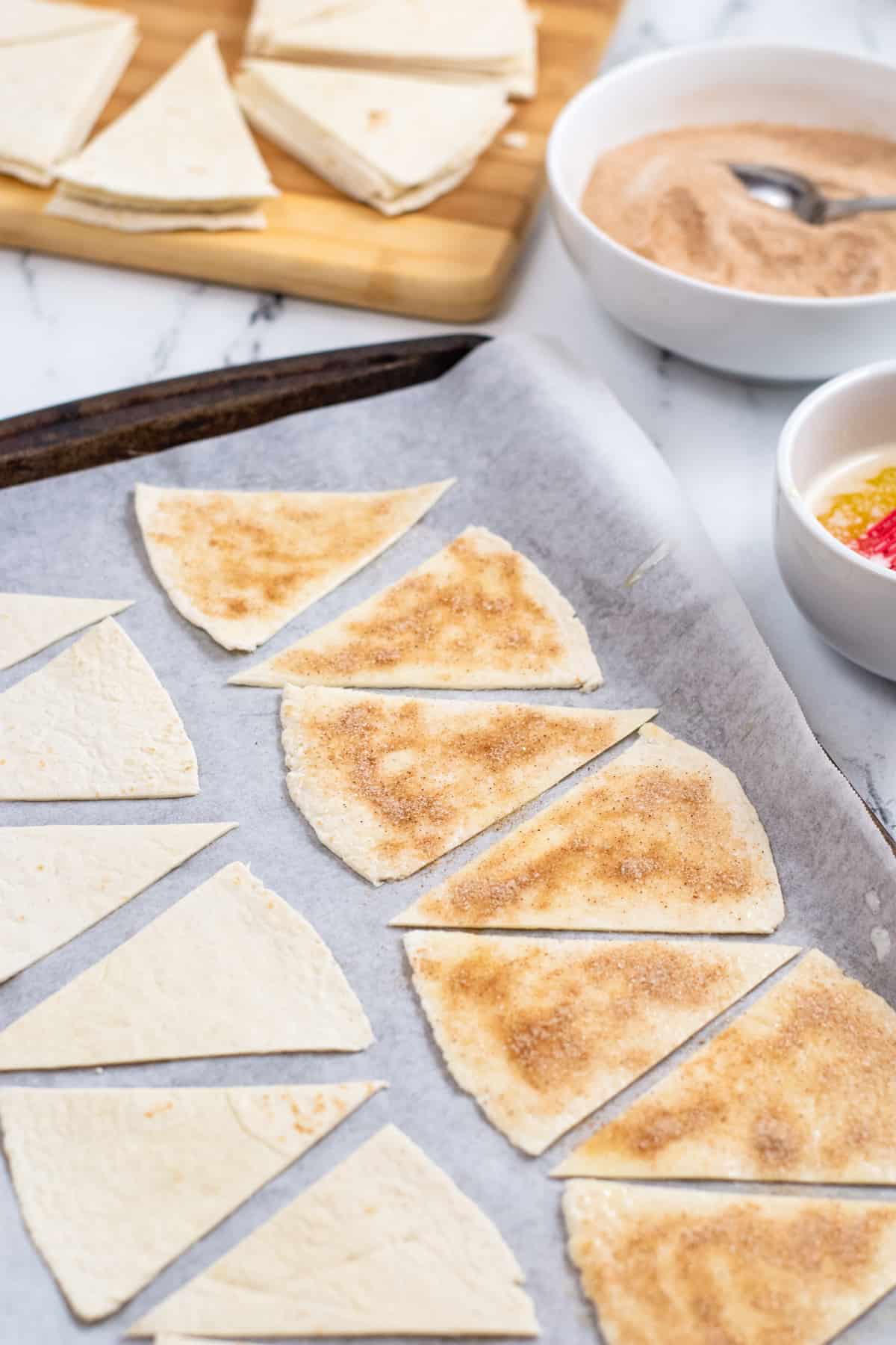 Triangular pieces of flour tortilla on parchment-lined baking sheet. Half are covered in butter, cinnamon, and sugar while the others are still plain. Bowl of cinnamon sugar and more tortilla can be seen in the background.